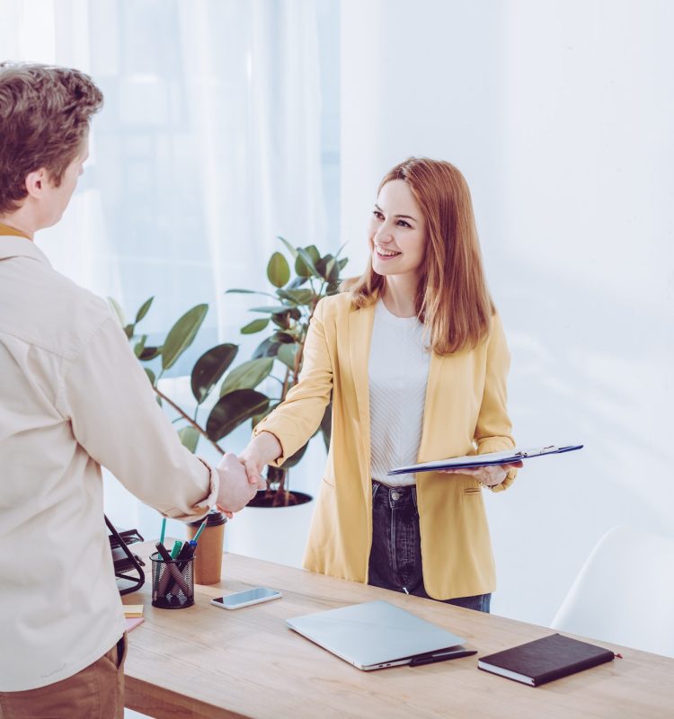 happy-recruiter-holding-clipboard-and-shaking-hands-with-employee-in-modern-office.jpg